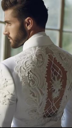 a man wearing a white suit with an intricate design on the back and shoulder, looking out a window