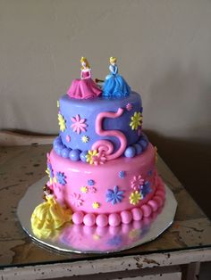 a pink and blue cake with princess figures on it sitting on a table next to a wall