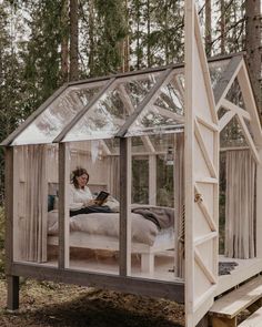 a woman sitting on a bed inside of a small glass house in the middle of a forest