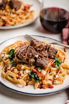 two plates with pasta and meat on them next to a glass of red wine in the background