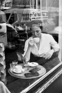 a woman sitting at a table in front of a store window drinking from a cup