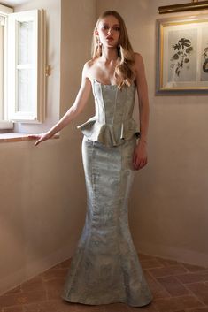 a woman in a silver dress standing by a window with her hand on the wall