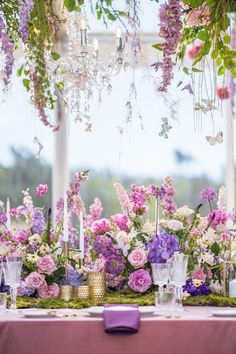 a table topped with lots of purple and white flowers next to tall vases filled with greenery