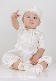 a baby sitting on top of a white table wearing a white dress and hat with one hand up in the air