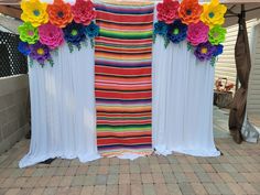 an outdoor area with white curtains and colorful flowers on the top, along with brick pavers flooring