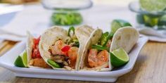 three shrimp tacos on a white plate with limes and water in the background