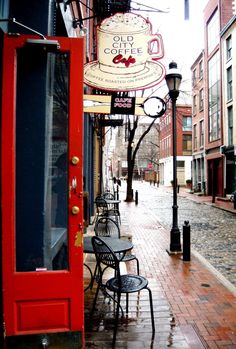 an empty sidewalk with tables and chairs next to a red door on the side walk