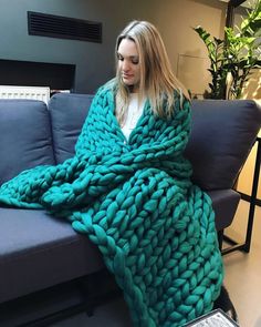 a woman is sitting on a couch wrapped in a large chunky knitted blanket