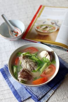 a bowl of soup with meat and vegetables on a table next to an open book