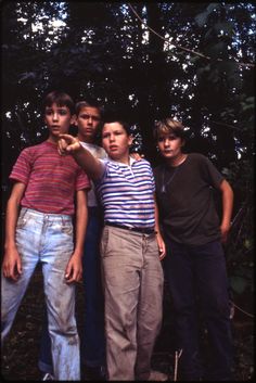 four young men posing for a photo in front of some trees and bushes, with one pointing at the camera