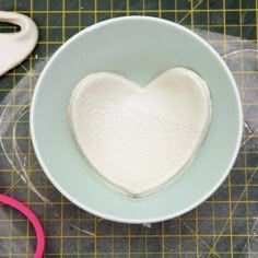 a white heart shaped bowl sitting on top of a table next to some pink scissors