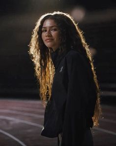 a woman with long curly hair standing in front of a track and looking off into the distance