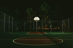 an outdoor basketball court at night with a basket ball in the air and trees behind it