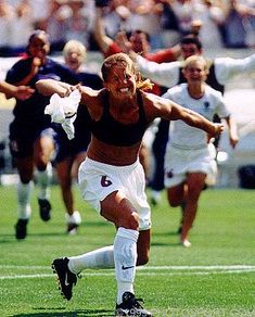 a woman kicking a soccer ball on top of a green field with other people watching