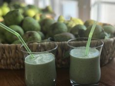 two glasses filled with green smoothie sitting on top of a table next to a basket of fruit