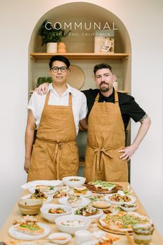 two men in aprons standing next to a table full of plates and bowls with food on it