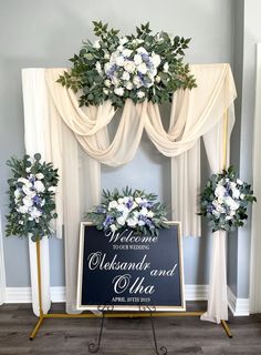 a welcome sign with flowers and greenery on it in front of a white drape