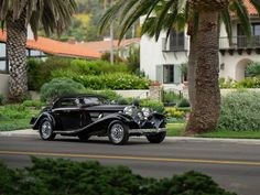 an old black car driving down the street in front of palm trees and houses with orange roofs