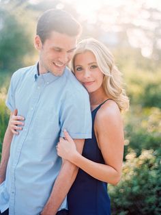 a man and woman hugging each other in front of some bushes with the sun shining on them