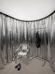 an empty chair and coat rack in front of a silver curtained room with shoes on the floor