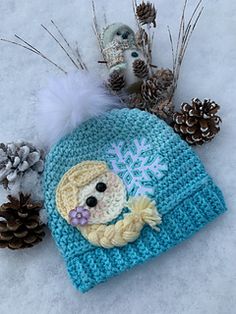 a crocheted hat with a little mermaid on it and pine cones in the snow