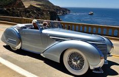 an old silver car driving down the road next to the ocean and cliff side area