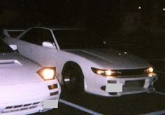 two white cars parked next to each other in a parking lot at night with lights on