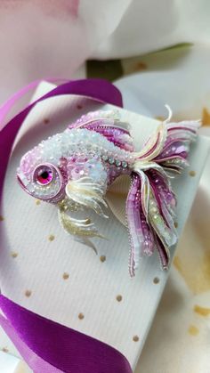 a pink and white fish brooch sitting on top of a piece of paper with purple ribbon