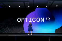 a woman standing on stage in front of a large screen that says opticon 19