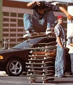 a man on a skateboard jumping over a stack of skateboards in front of a car