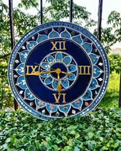 a blue and gold clock sitting on top of green bushes in front of a fence