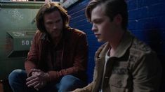 two young men sitting next to each other in front of a blue wall and looking at something