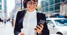 a woman in glasses holding a cup and looking at her cell phone while walking down the street