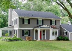 a large gray house with black shutters on the front and red door is surrounded by trees