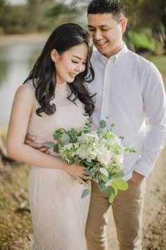 a young man and woman standing next to each other holding flowers in front of a body of water