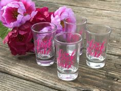 three shot glasses sitting on top of a wooden table next to pink flowers and greenery