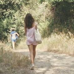 two people walking down a dirt road with trees in the background and bushes on either side