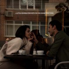 a man and woman sitting at a table with cups in front of them, talking