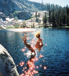 two women in bikinis jumping into the water from rocks near a lake with pine trees