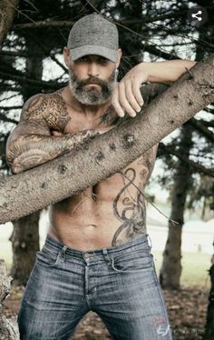 a shirtless man with tattoos on his chest and arms leaning against a tree branch