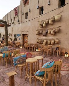 an outdoor seating area with wooden tables and chairs in front of a tan wall that has hanging baskets on it