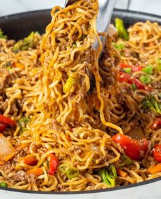 a skillet filled with noodles, meat and veggies being stirred by tongs