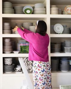 a woman in pink shirt and white pants reaching up to shelves with plates on them