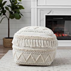 a white poufce with tassels sits on the floor in front of a fireplace