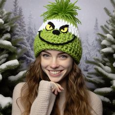 a woman wearing a knitted hat with a green monster face on it, in front of snow - covered trees