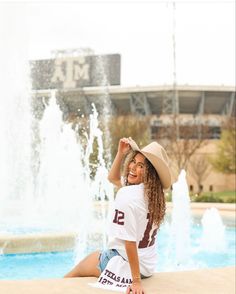 a woman sitting in front of a fountain wearing a cowboy hat and white t - shirt