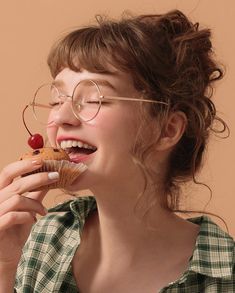 a woman wearing glasses eating a cupcake with cherries on her nose and smiling