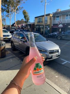 a person holding up a pink drink in front of a parked car on the street