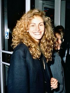 a woman with curly hair standing in front of a window and smiling at the camera