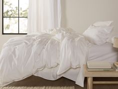 an unmade bed with white sheets and pillows in front of a window on a wooden floor